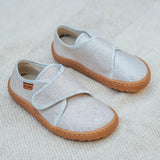 Froddo barefoot kids slipper in gold shine available at Babipur standing on a soft pale blanket. A canvas style slipper shoe in a light neutral colour with a velcro fastener for a secure fit and a dark brown grippy sole