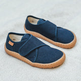 Froddo barefoot kids slipper in dark blue available at Babipur standing on a soft pale blanket. A canvas style slipper shoe in a light neutral colour with a velcro fastener for a secure fit and a dark brown grippy sole