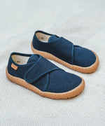 Froddo barefoot kids slipper in dark blue available at Babipur standing on a soft pale blanket. A canvas style slipper shoe in a light neutral colour with a velcro fastener for a secure fit and a dark brown grippy sole