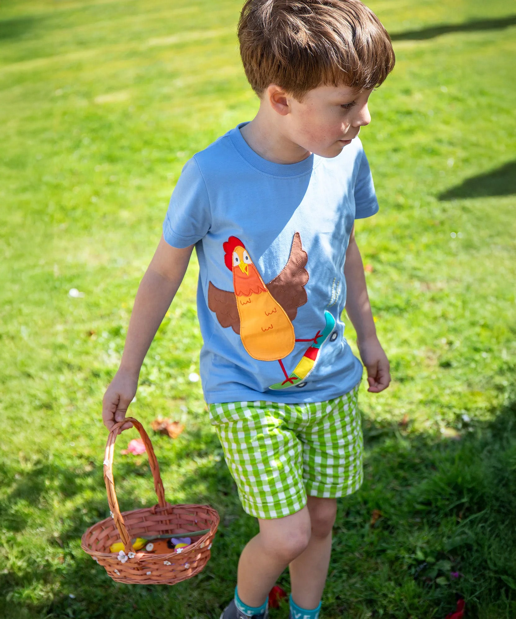 A child wearing the Frugi Avery appliqué t-shirt, a soft GOTS organic cotton light blue top with a chicken riding a skateboard appliqué on the front.