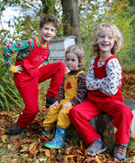 Three children sitting on a large tree stump. Two are wearing the red Frugi Babipur cord dungarees and one is wearing teh yellow version.