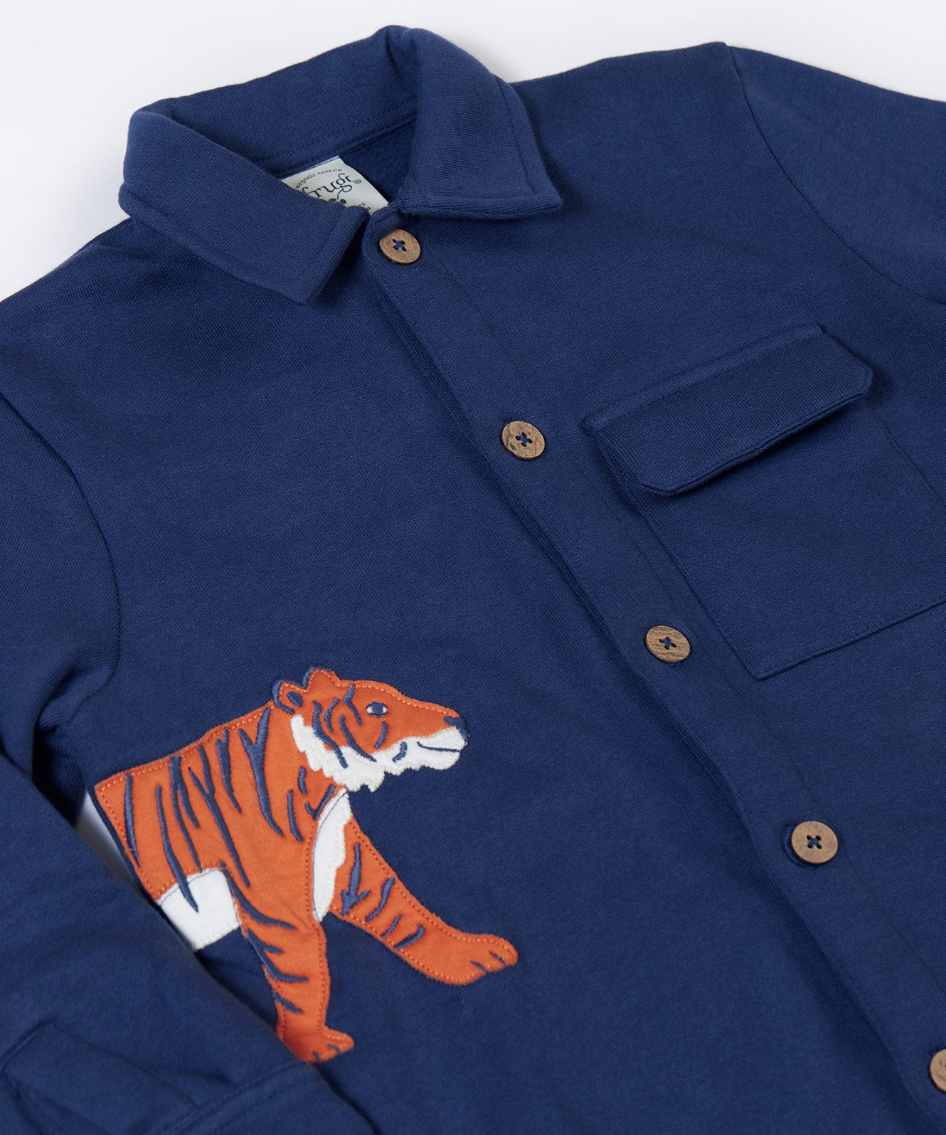 A close up of the orange tiger applique on the side of the Frugi Big Cats Navy Organic Cotton Cosy Kids' Shirt Shaket. This shaket has a tiger and cheetah applique details on the sides. This navy shirt jacket is a part of a wide range of organic cotton clothing for children available here at Babipur.