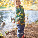 A toddler standing by a lake and wearing the Frugi puddle buster navy trousers, be hoppy coat and matching puddle buster wellington boots  A natural rubber and waterproof wellington boot in navy with red trim with prints of multicoloured frogs.