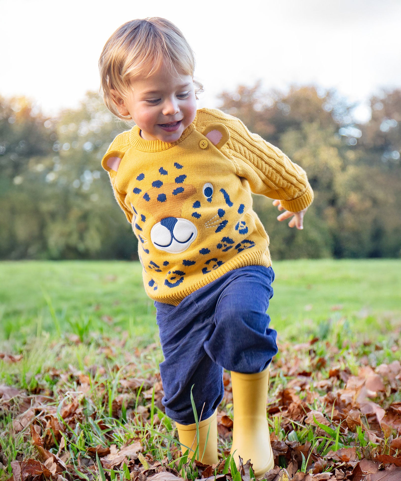 A child walking on grass wearing the Frugi Caleb Leopard Character Knit Jumper, navy cord trousers and yellow wellington boots. This knitted jumper is a part of a wide range of organic cotton clothing for children available here at Babipur.