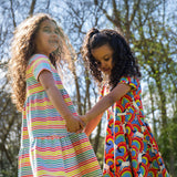 Two children wearing Frugi organic cotton dresses. One is wearing the Cornflower Rainbow Stripe Lucy dress the other is wearing the Over The Rainbow Skater dress .