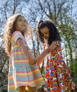 Two children wearing Frugi organic cotton dresses. One is wearing the Cornflower Rainbow Stripe Lucy dress the other is wearing the Over The Rainbow Skater dress .