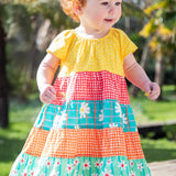 A toddler wearing the Frugi Dorothy Twirly Dress, with a Rainbow Hotchpotch print, a patchwork type design featuring yellow, red orange and green patterns with daisies, stood amongst trees.