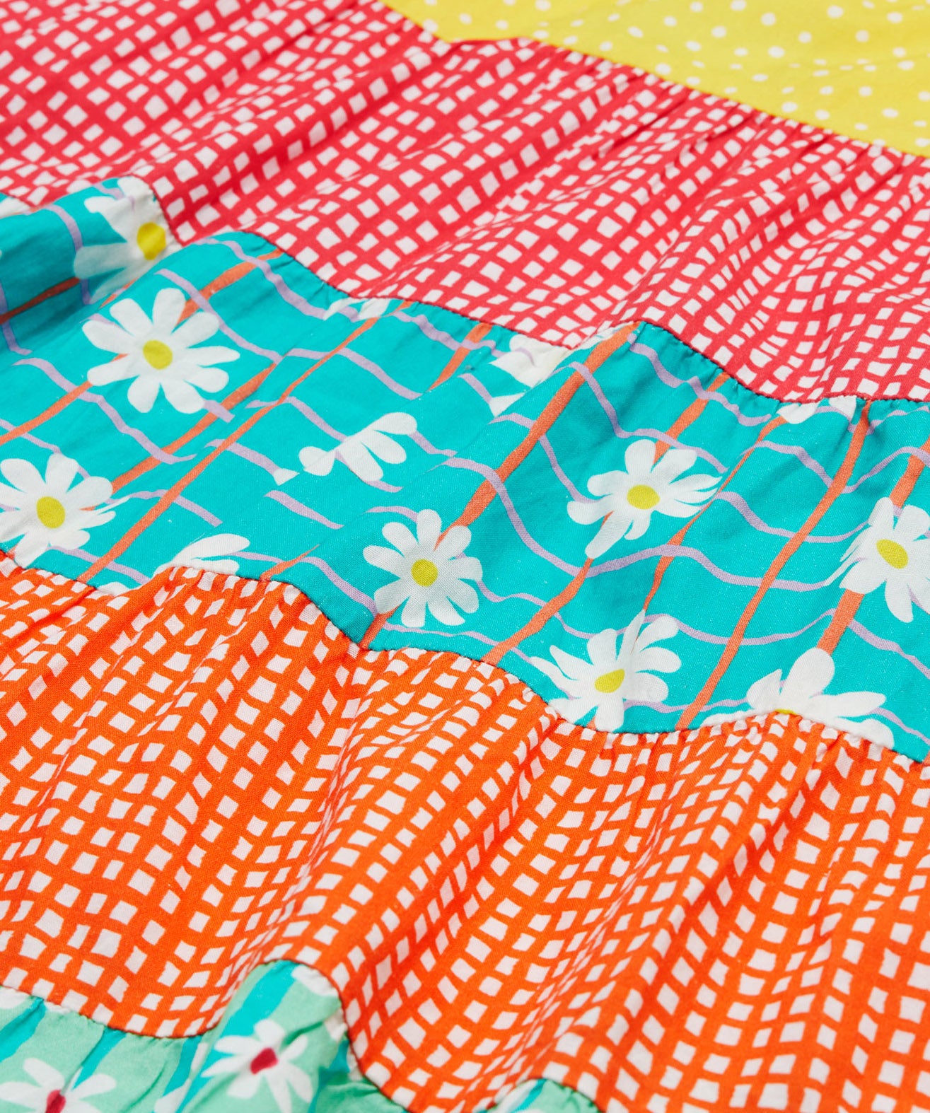 A close up of Frugi Dorothy Twirly Dress, with a Rainbow Hotchpotch print, a patchwork type design featuring orange, mint, red, yellow and green patterns with daisies.