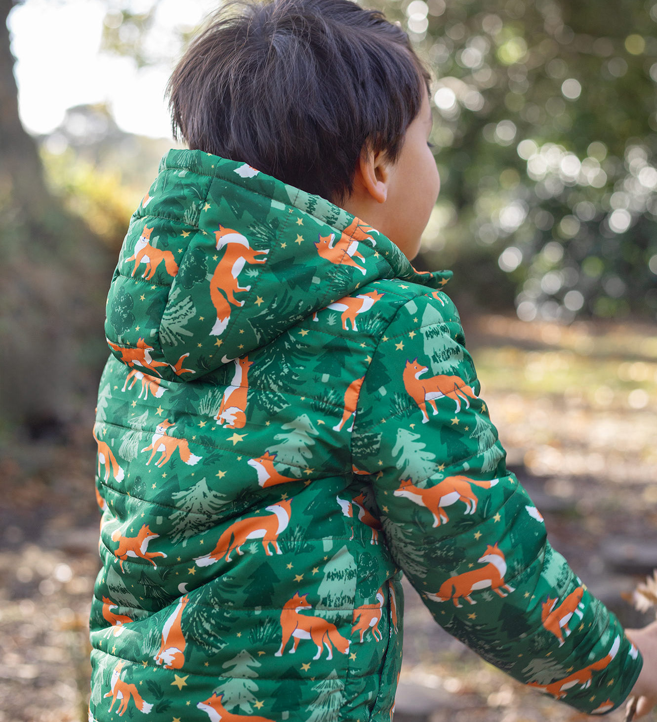 A child wearing the Frugi toasty trail reversible jacket showing the playful fox in woodland design and hood detail on green post consumer recycled polyester fabric