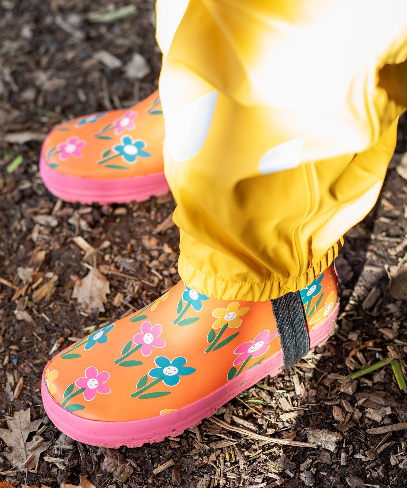 A child standing outside and wearing the Frugi puddle buster yellow trousers and puddle buster wellington boots in fun flowers  A natural rubber and waterproof wellington boot in orange with prints of multicoloured, playful flowers.