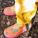 A child standing outside and wearing the Frugi puddle buster yellow trousers and puddle buster wellington boots in fun flowers  A natural rubber and waterproof wellington boot in orange with prints of multicoloured, playful flowers.