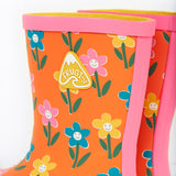 A close up of the top of the Frugi kids puddle buster wellington boots. Showing the the pink trim and flower print detail on orange natural rubber material. Features a yellow triangular brand patch on the outside of the boot.