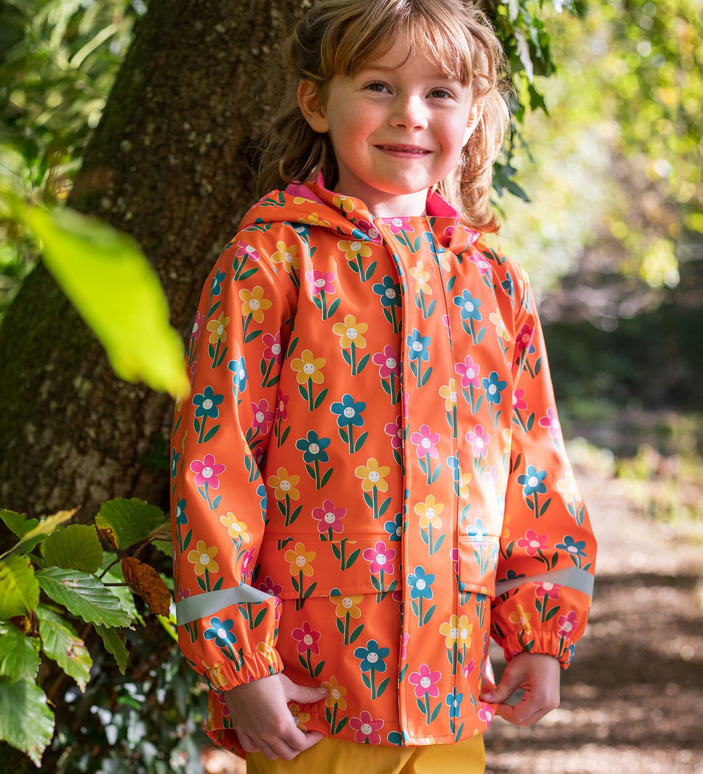 A toddler standing by a tree and wearing the Frugi puddle buster yellow trousers, fun flowers coat.  An orange waterproof coat with playful prints of flowers.