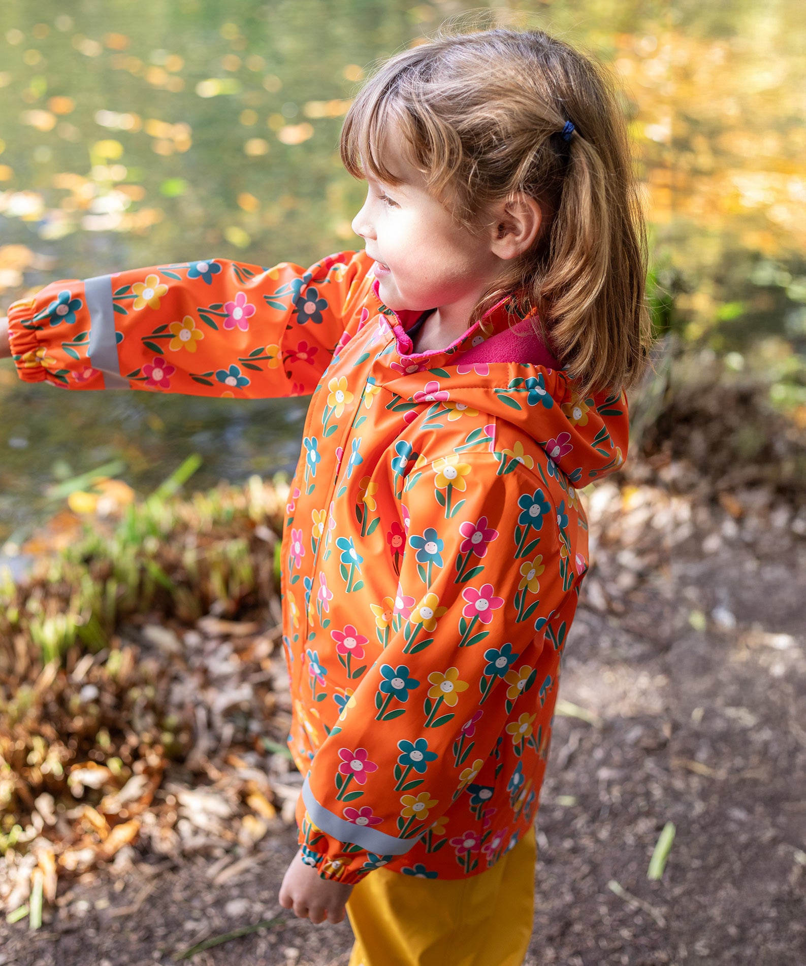 A side view of a toddler wearing the Frugi fun flowers puddle buster coat showing the hood detail and pink fleece inside liner.