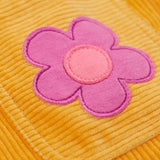 A close up of a pink flower on the Frugi organic cotton Ginny yellow gold Corduroy Pinafore Dress with Flowers with retro flower tights and a rainbow striped top underneath. This dress is a part of a wide range of organic cotton clothing for children available here at Babipur.