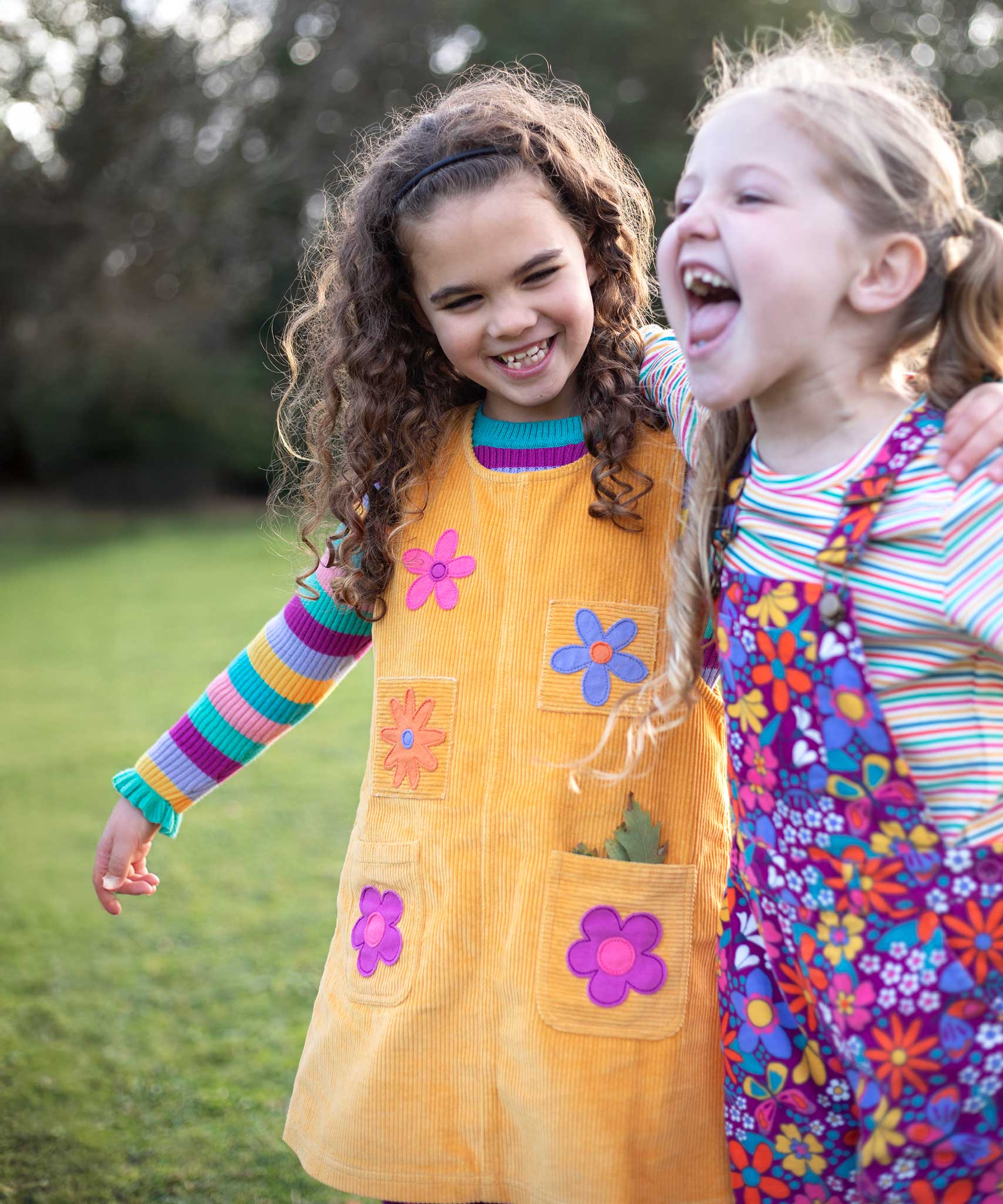 A child wearing the Frugi organic cotton Ginny yellow gold Corduroy Pinafore Dress with Flowers with retro  flower tights and a rainbow striped top underneath. This dress is a part of a wide range of organic cotton clothing for children available here at Babipur.