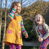 A child wearing the Frugi organic cotton Ginny yellow gold Corduroy Pinafore Dress with Flowers with retro  flower tights and a rainbow striped top underneath. This dress is a part of a wide range of organic cotton clothing for children available here at Babipur.