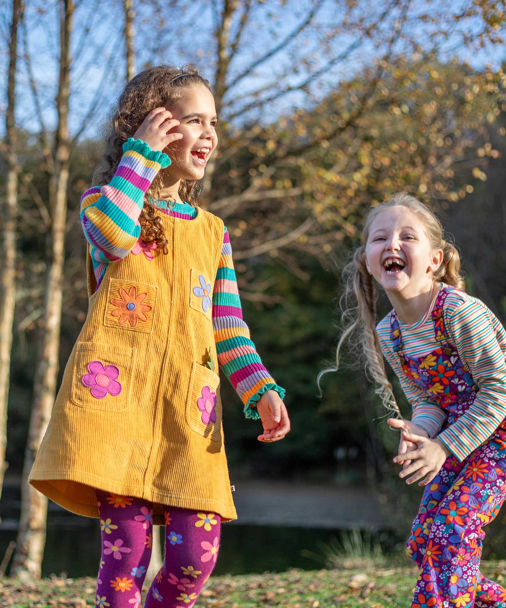 A child wearing the Frugi organic cotton Ginny yellow gold Corduroy Pinafore Dress with Flowers with retro  flower tights and a rainbow striped top underneath. This dress is a part of a wide range of organic cotton clothing for children available here at Babipur.