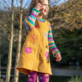 A child wearing the Frugi organic cotton Ginny yellow gold Corduroy Pinafore Dress with Flowers with retro  flower tights and a rainbow striped top underneath. This dress is a part of a wide range of organic cotton clothing for children available here at Babipur.