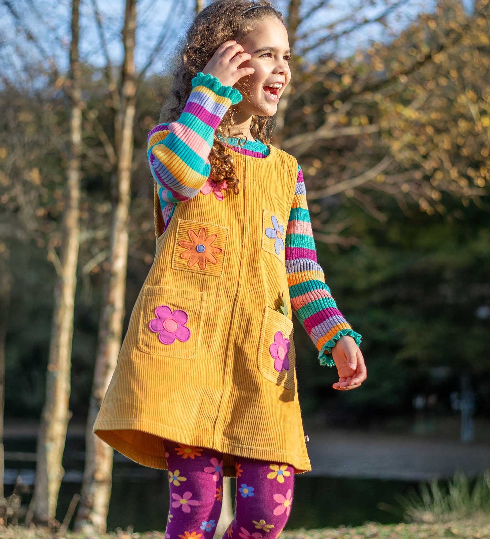 A child wearing the Frugi organic cotton Ginny yellow gold Corduroy Pinafore Dress with Flowers with retro  flower tights and a rainbow striped top underneath. This dress is a part of a wide range of organic cotton clothing for children available here at Babipur.