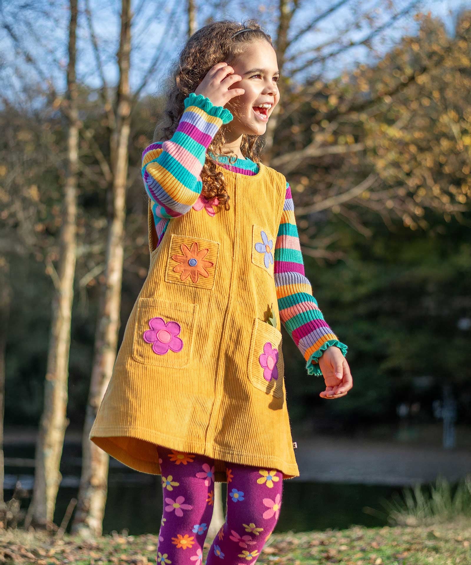 A child wearing the Frugi organic cotton Ginny yellow gold Corduroy Pinafore Dress with Flowers with retro  flower tights and a rainbow striped top underneath. This dress is a part of a wide range of organic cotton clothing for children available here at Babipur.