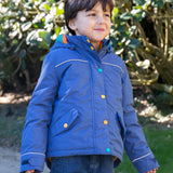 A child wearing the Frugi autumn stripe /navy waterproof rambler coat. A thick and warm post consumer recycled polyester coat in navy with multicoloured popper fasteners.