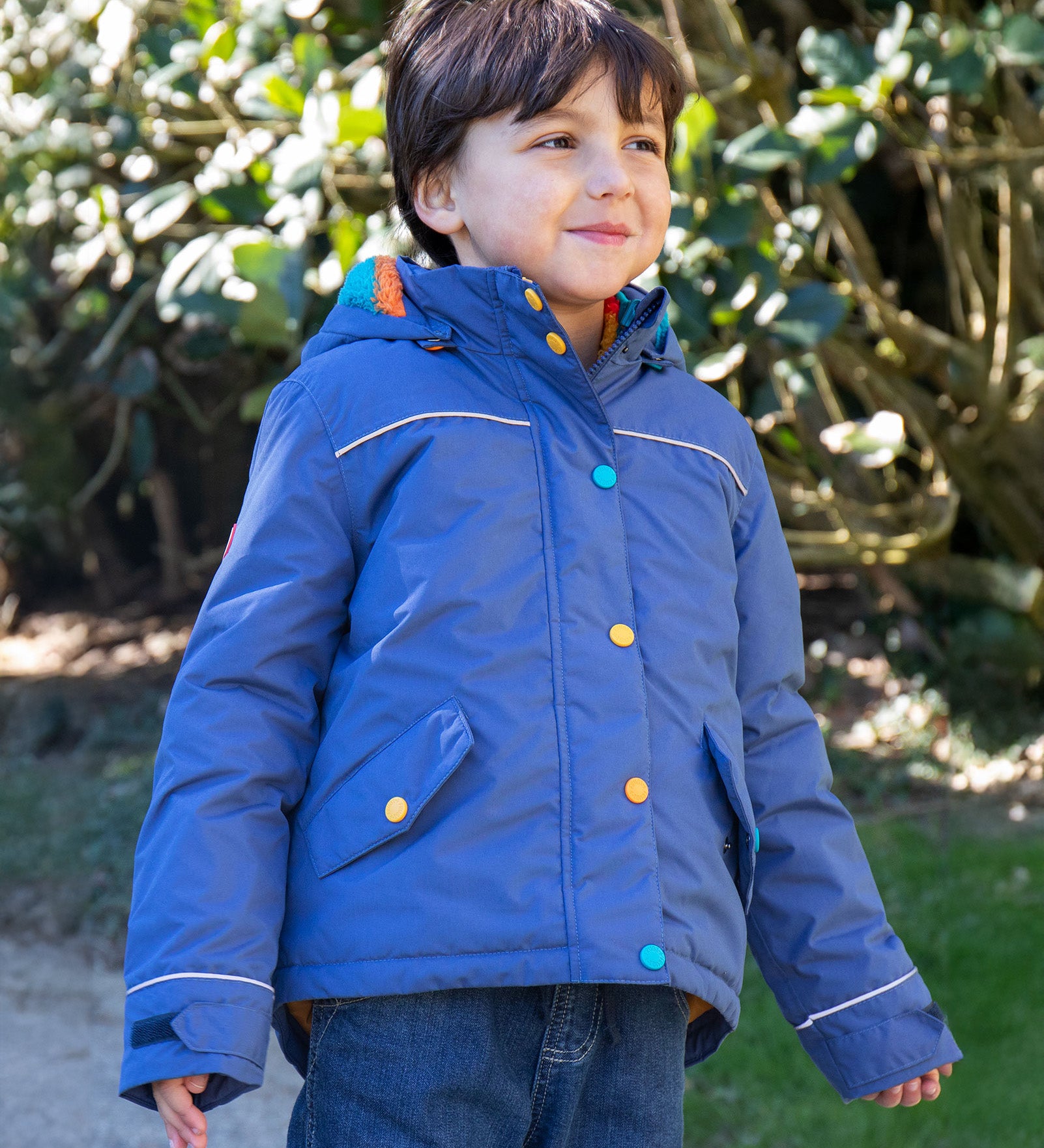 A child wearing the Frugi autumn stripe /navy waterproof rambler coat. A thick and warm post consumer recycled polyester coat in navy with multicoloured popper fasteners.