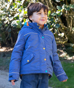 A child wearing the Frugi autumn stripe /navy waterproof rambler coat. A thick and warm post consumer recycled polyester coat in navy with multicoloured popper fasteners.