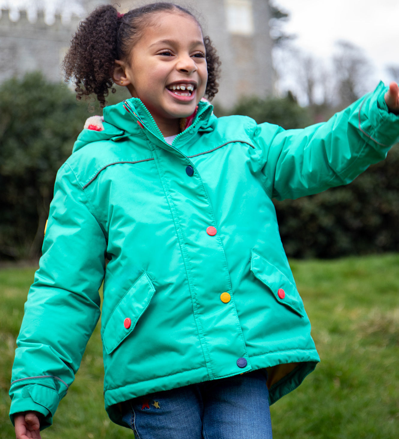 A child wearing the Frugi dragon/mushroom waterproof rambler coat. A thick and warm post consumer recycled polyester coat in bright turquoise with multicoloured popper fasteners.