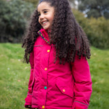A child wearing the Frugi snapdragon/navy waterproof rambler coat. A post consumer recycled polyester coat in vibrant pink with multicoloured popper fasteners.