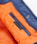 A close up of the Frugi kids 3 in 1 rambler coat in Autumn stripe /navy colourway showing the hidden side pocket and navy zip detail on the inside of the coat on orange post consumer recycled polyester fabric