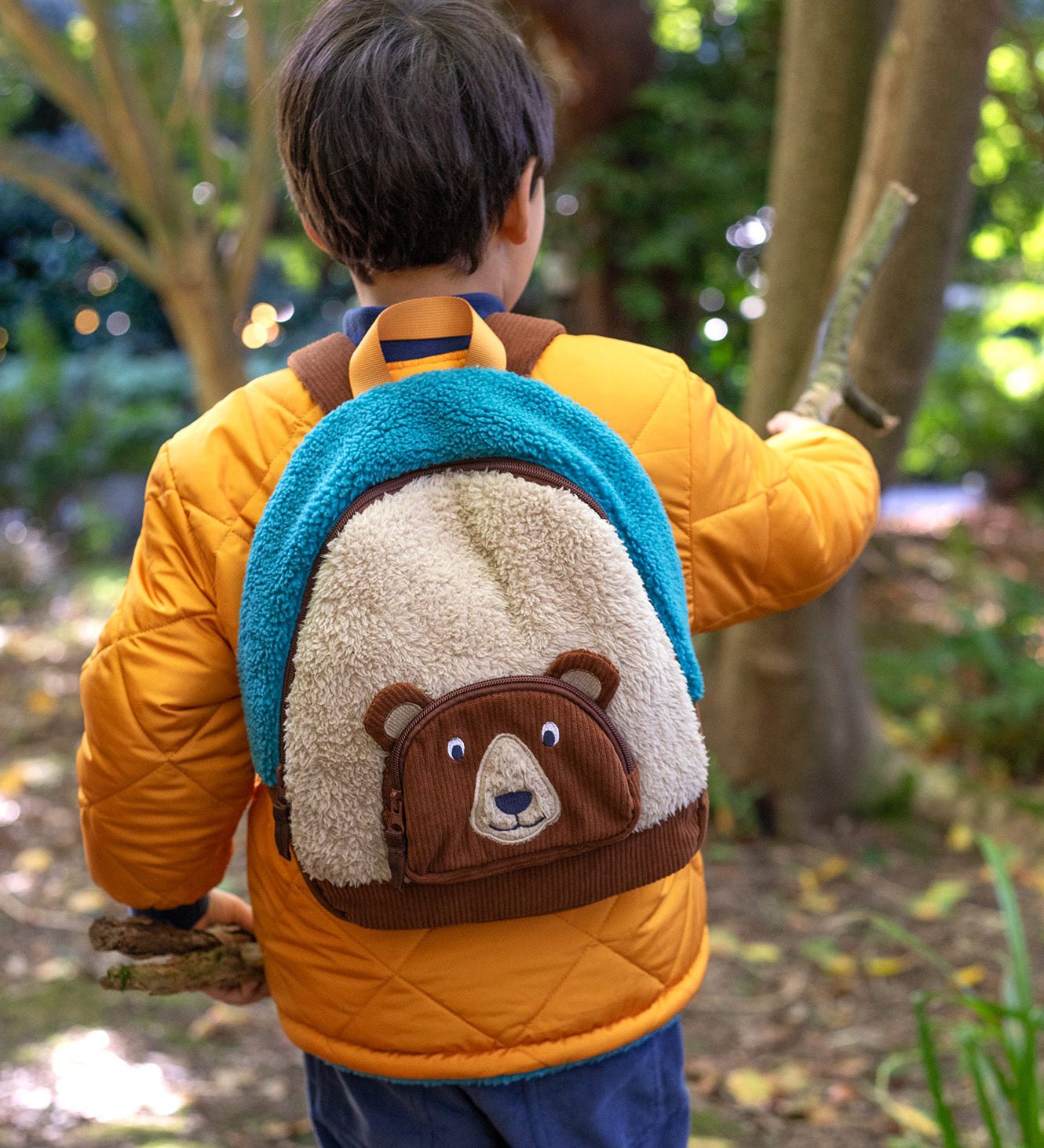 A child wearing the Frugi kids beary wild backpack outside showing scale. A blue and neutral colour fleece type with a corduroy character bear as a front pocket. Features a handy yellow carry handle on the top and brown corduroy shoulder straps