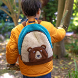 A child wearing the Frugi kids beary wild backpack outside showing scale. A blue and neutral colour fleece type with a corduroy character bear as a front pocket. Features a handy yellow carry handle on the top and brown corduroy shoulder straps