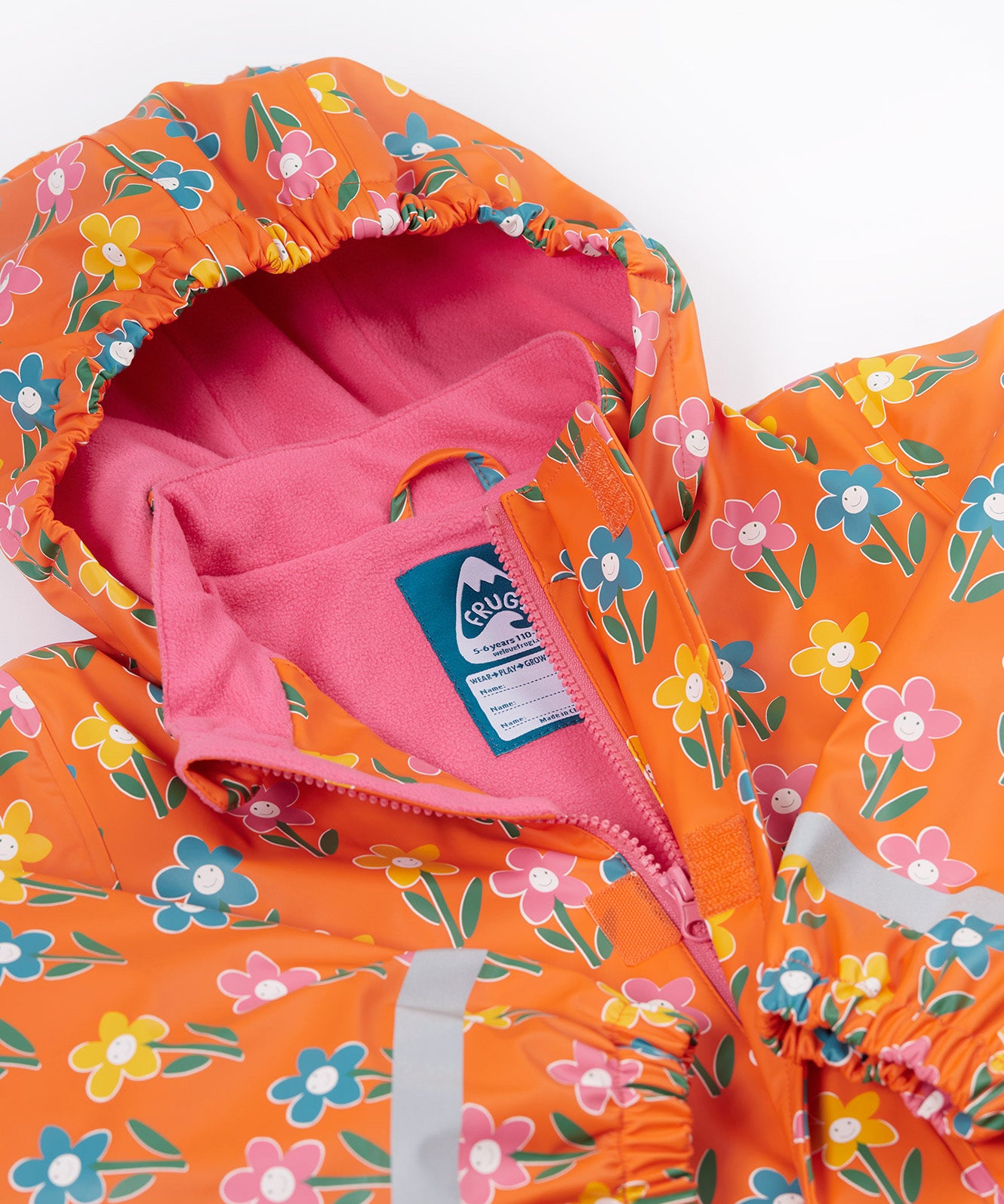 A close up of the Frugi kids fun flowers puddle buster coat showing the multicolour flowers on orange waterproof material. Features a full length zip and velcro fasteners for a secure fit.