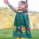 A child standing outside and wearing the Frugi kids pippa holly green fun guys pinafore outfit. A 100% GOTS organic cotton woodland inspired outfit which features a pink long sleeve top and gree n pinafore