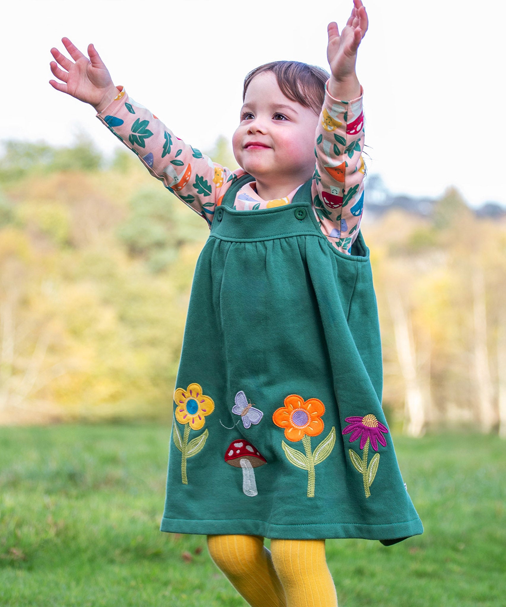 A child standing outside and wearing the Frugi kids pippa holly green fun guys pinafore outfit. A 100% GOTS organic cotton woodland inspired outfit which features a pink long sleeve top and gree n pinafore