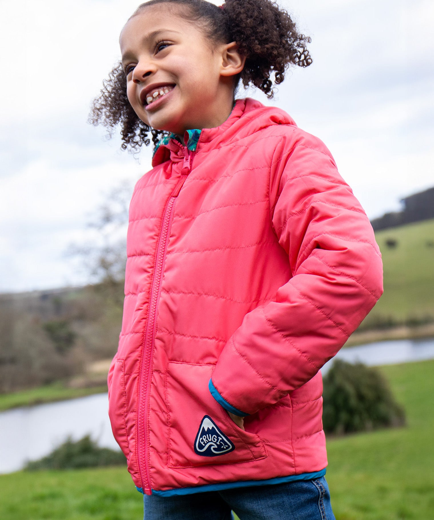 A child wearing the Frugi toasty trail reversible jacket showing the light pink coloured with pink zip detail and a brand patch on the side pocket on post consumer recycled polyester fabric.