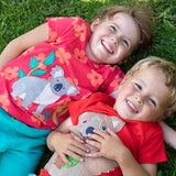 Children lying on the grass. One child wearing the Frugi Carsen t-shirt - a red with short sleeves and a playful appliqué of a brown quokka. Frugi Lottie t-shirt - a watermelon pink with short sleeve and an appliqué of a grey koala