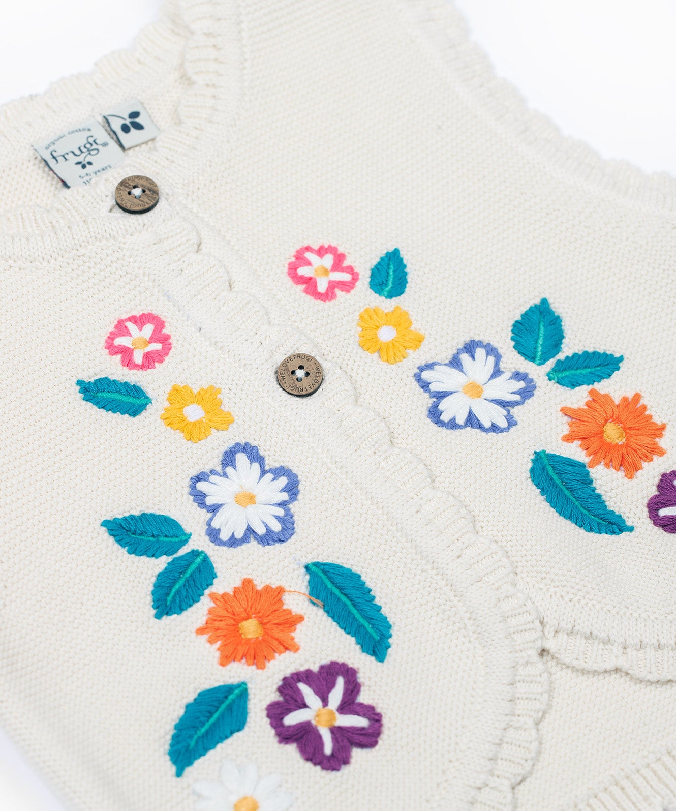 A close up of the embroidered flowers on the front of the Frugi Oatmeal Flowers Connie Cotton Knitted Sleeveless Cardigan. This white oatmeal sleeveless cardigan has cute scalloped edges and is decorated with pretty embroidered flowers and leaves on the front.  This waistcoat is a part of a wide range of organic cotton clothing for children available here at Babipur.