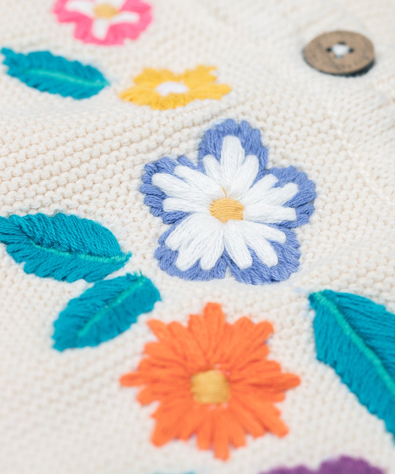 A close up of one of the embroidered flowers on the front of the Frugi Oatmeal Flowers Connie Cotton Knitted Sleeveless Cardigan. This white oatmeal sleeveless cardigan has cute scalloped edges and is decorated with pretty embroidered flowers and leaves on the front.  This waistcoat is a part of a wide range of organic cotton clothing for children available here at Babipur.