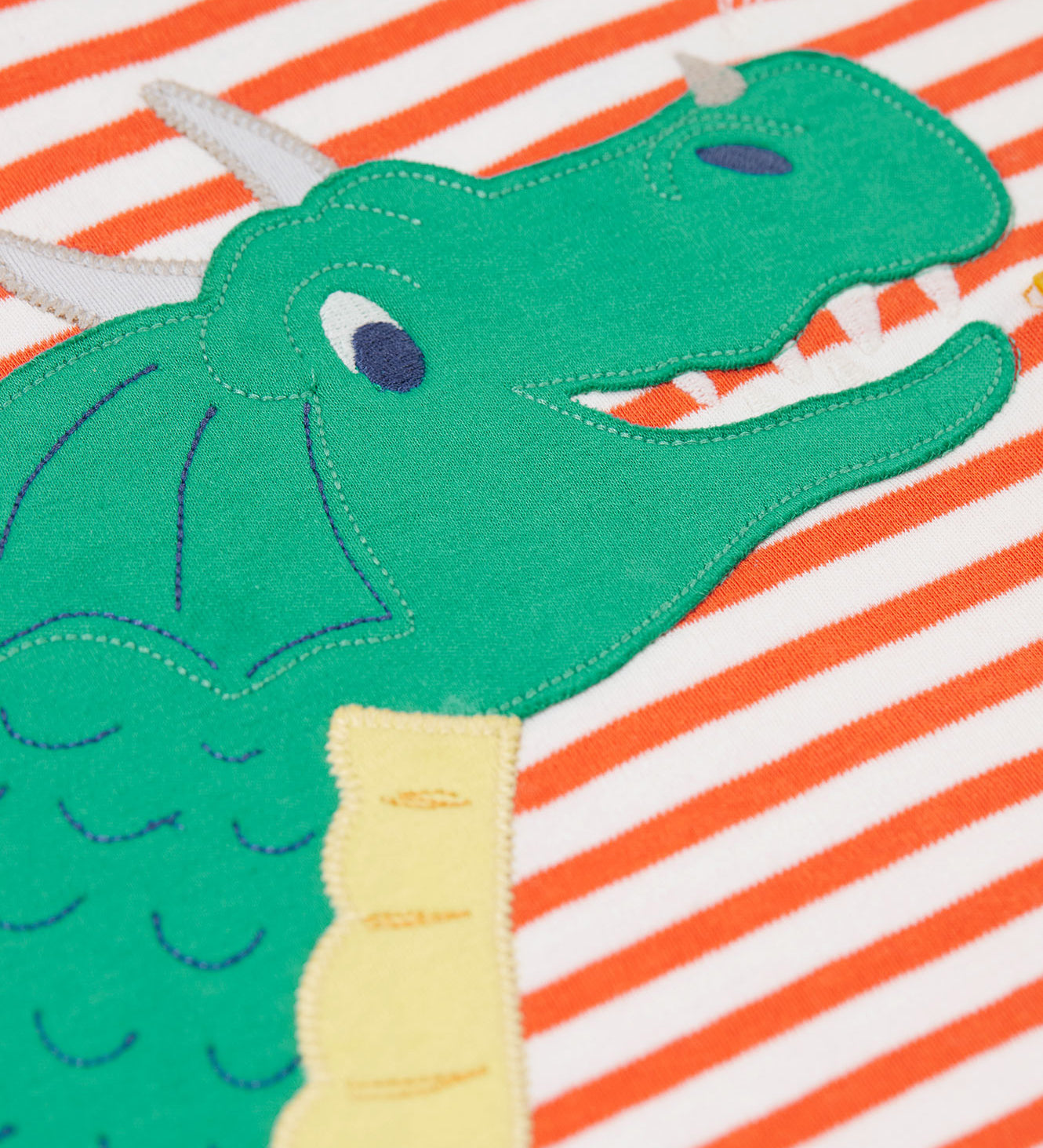 A close up of the dragon's face on the front of the Frugi Salamander Stripe Dragon Discovery Circular Top. This orange and white striped top has a green coloured dragon applique detail on the front. This top is a part of a wide range of organic cotton clothing for children available here at Babipur.