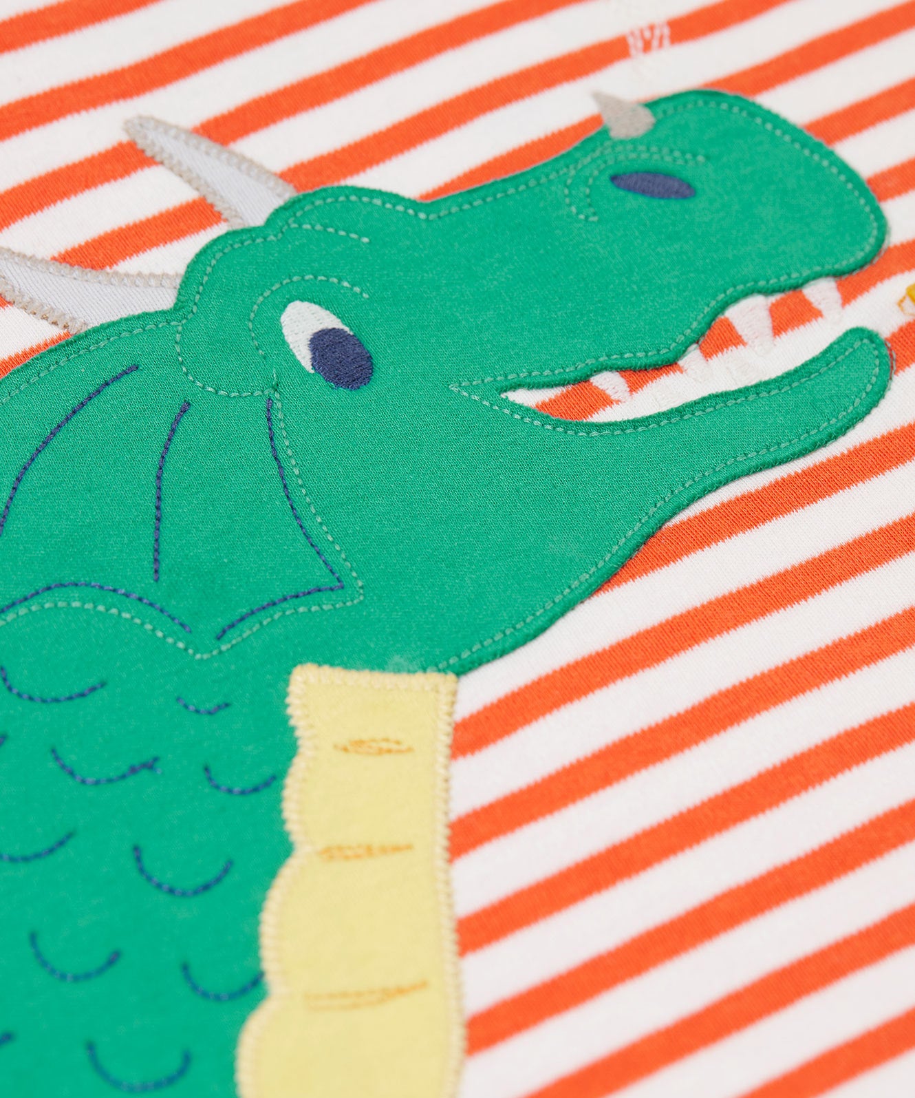 A close up of the dragon's face on the front of the Frugi Salamander Stripe Dragon Discovery Circular Top. This orange and white striped top has a green coloured dragon applique detail on the front. This top is a part of a wide range of organic cotton clothing for children available here at Babipur.