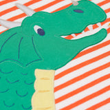 A close up of the dragon's face on the front of the Frugi Salamander Stripe Dragon Discovery Circular Top. This orange and white striped top has a green coloured dragon applique detail on the front. This top is a part of a wide range of organic cotton clothing for children available here at Babipur.