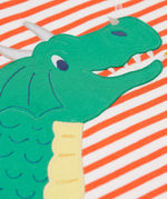 A close up of the dragon's face on the front of the Frugi Salamander Stripe Dragon Discovery Circular Top. This orange and white striped top has a green coloured dragon applique detail on the front. This top is a part of a wide range of organic cotton clothing for children available here at Babipur.