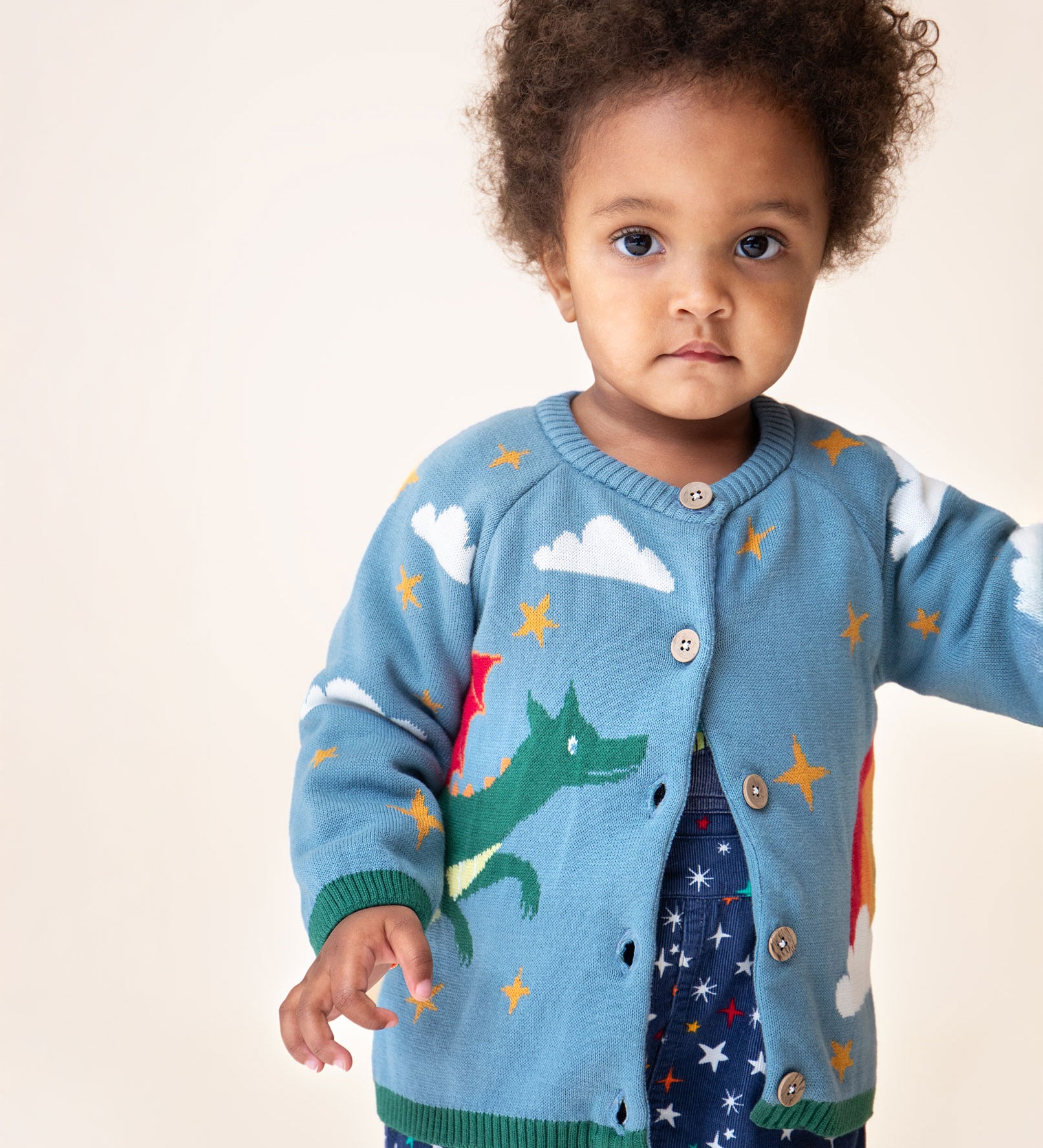 A child wearing the Frugi Organic Cotton Reva Dragon Skies Knitted Kids' Cardigan. This blue cardigan features a fun, colourful design featuring a green dragon, stars, rainbow, clouds and a mushroom. This cardigan is a part of a wide range of organic cotton clothing for children available here at Babipur.