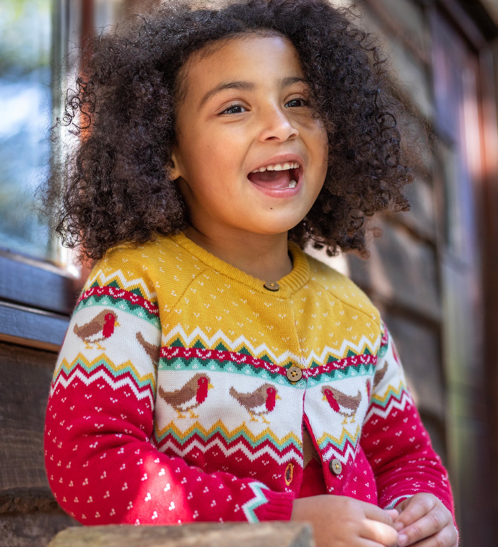 A child wearing the Frugi Festive Fair Isle Organic Cotton Kids' Carol Cardigan featuring red, yellow and green patterns and robin redbreast details. 
This knitted cardigan is a part of a wide range of organic cotton clothing for children available here at Babipur.