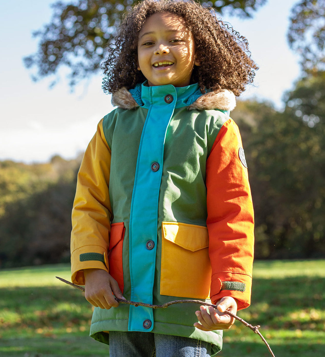 A child wearing the Frugi kids penhale Khaki parka outside- A long length thick warm coat  furry hood for warmth and  dark orange and yellow side pockets.