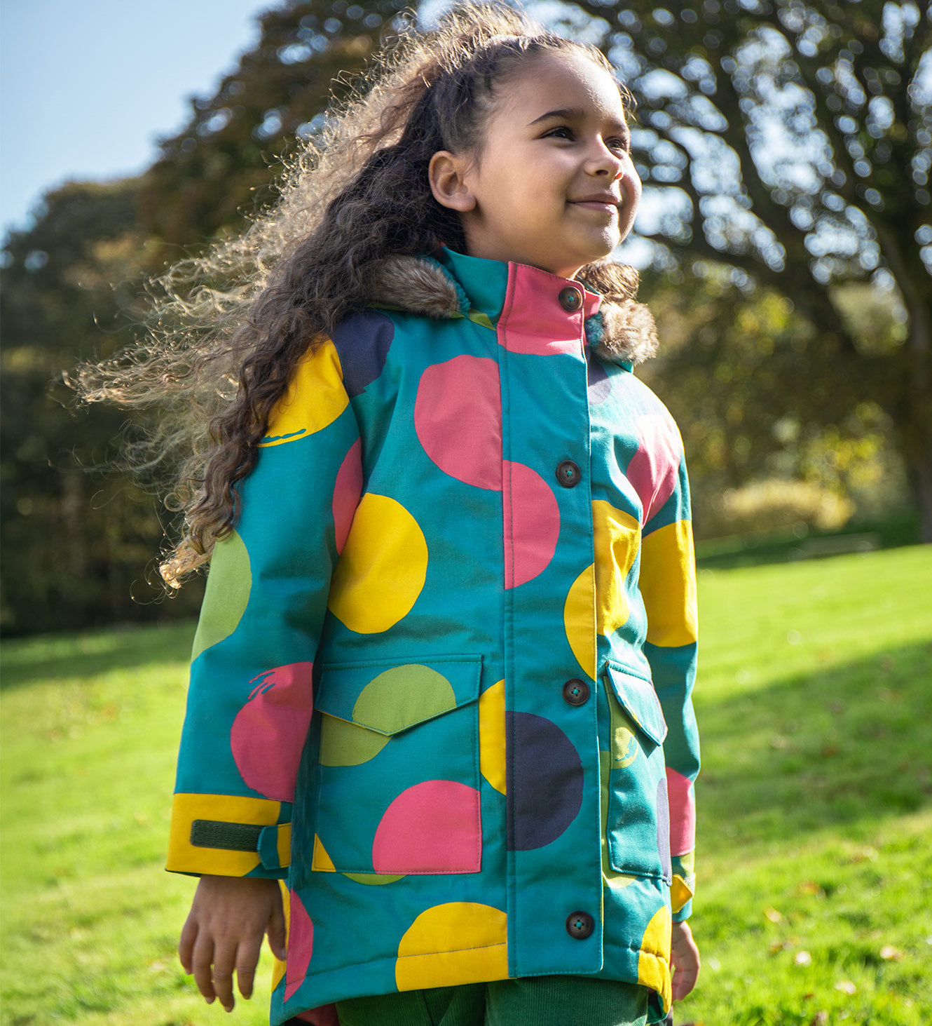 A child wearing the Frugi kids penhale super spot parka outside- A long length  organic cotton fabric warm coat with furry hood for warmth. Features a velcro strap on the arms to adjust the fit
