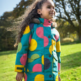 A child wearing the Frugi kids penhale super spot parka outside- A long length  organic cotton fabric warm coat with furry hood for warmth. Features a velcro strap on the arms to adjust the fit