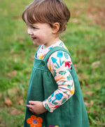 A child standing outside and wearing the Frugi kids pippa holly green fun guys pinafore outfit. A 100% GOTS organic cotton which features a pink long sleeve top and green pinafore with woodland inspired prints and appliques